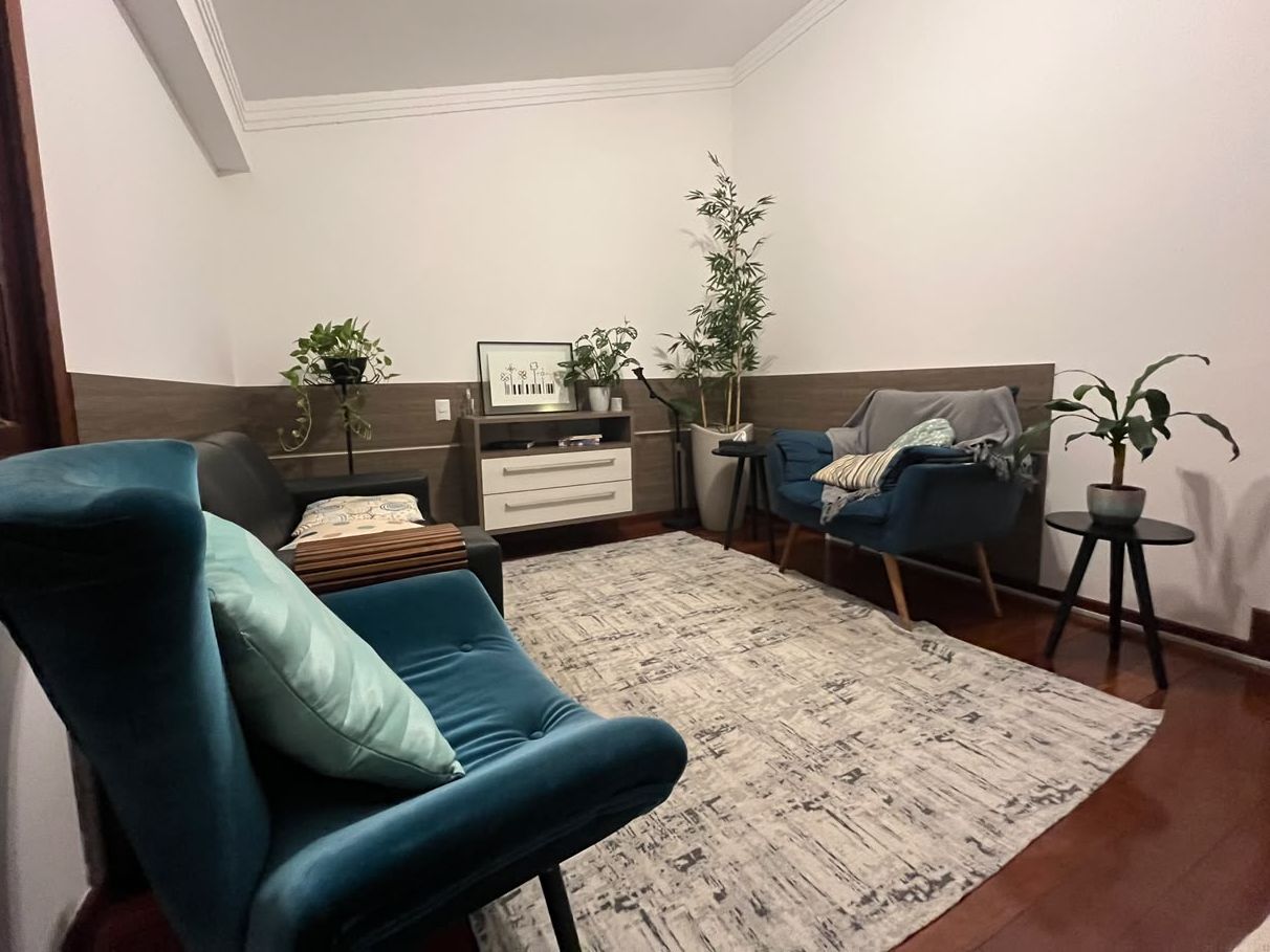 Outra vista do consultório, com poltronas azuis, tapete claro, plantas e uma cômoda ao fundo
